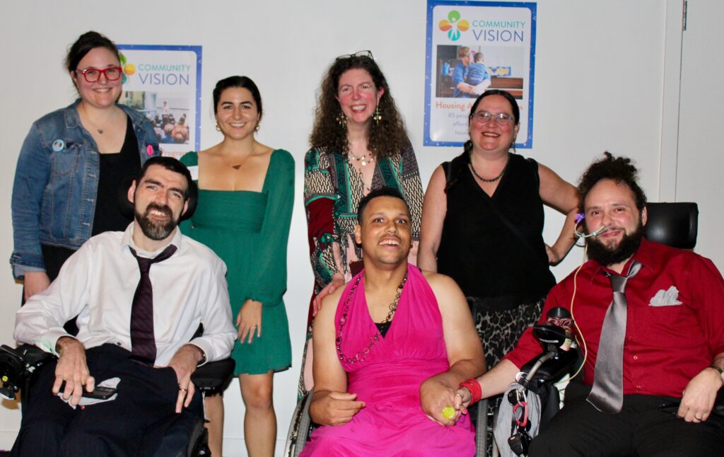 Seven smiling people who are dressed up for a party pose for a group photo, four of them are standing and three are seated in wheelchairs.
